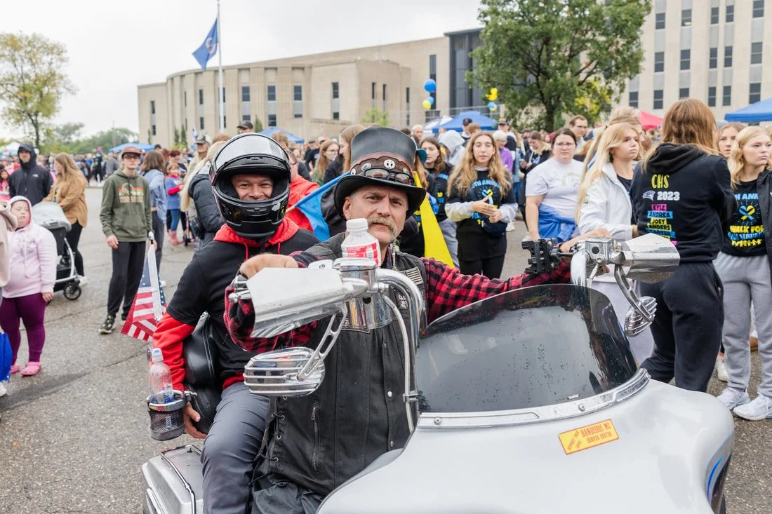 Two men on a motorcycle at a large outdoor gathering or protest, with many people and some flags in the background.