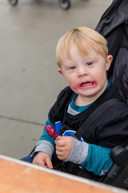 A young boy with red hair and light skin, sitting in a stroller, holding a pink ice cream popsicle, with chocolate and strawberry smeared around his mouth.