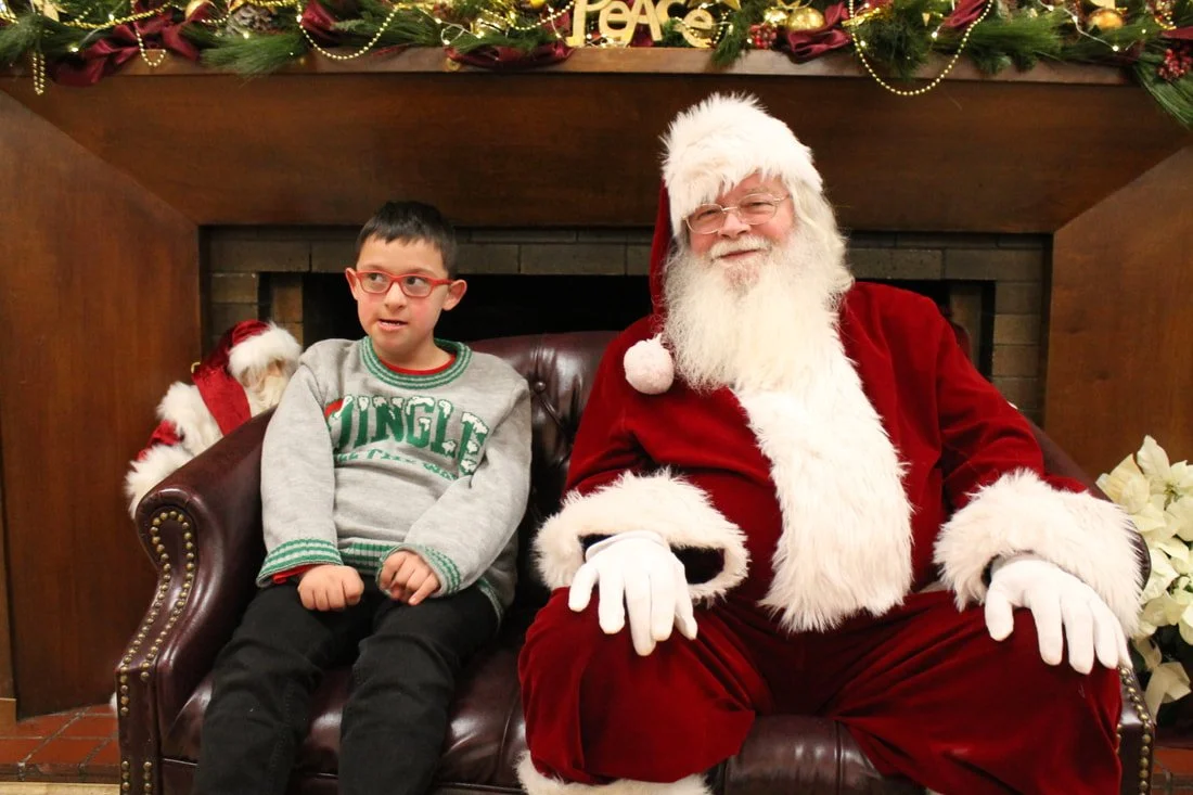 A young boy sitting next to Santa Claus on a leather couch, both smiling, in front of a decorated fireplace with Christmas ornaments and greenery.