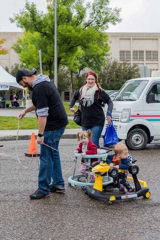 A man pulling a toddler in a wagon with a young girl sitting inside, and a boy sitting on a toy ride-on at a street event with tents and trees in the background.