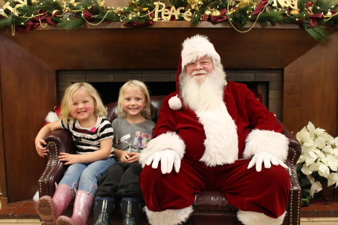 Two young girls sitting on a leather couch next to a man dressed as Santa Claus, all smiling. The scene is decorated with Christmas garland and ornaments, with a fireplace in the background.