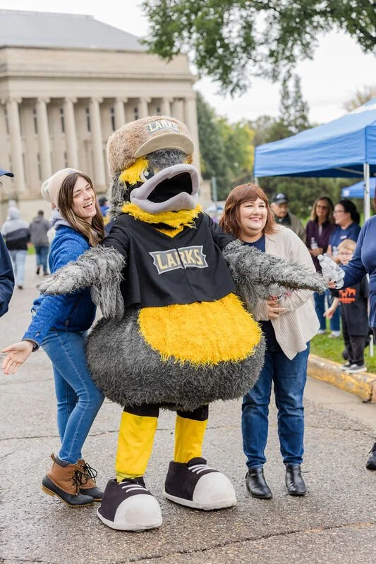 People with a large mascot dressed as a bird in a black shirt, yellow feathers, and sneakers, standing outdoors during a community event.