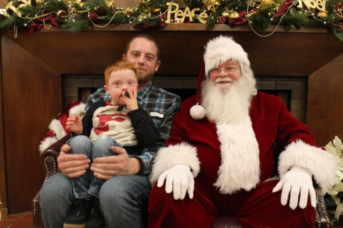 A man and a young boy sitting on a decorated Christmas bench with Santa Claus, in front of a festive garland and fireplace.