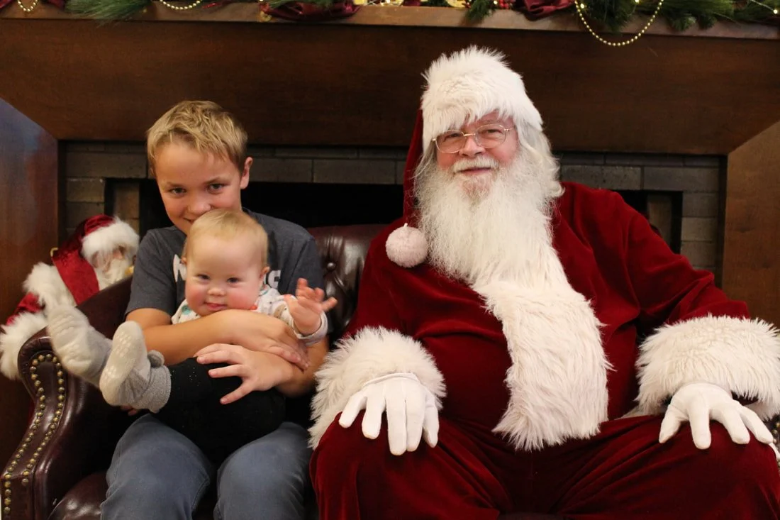A young boy and a baby sitting with Santa Claus on a holiday themed set, with Christmas decorations and a Christmas tree in the background.