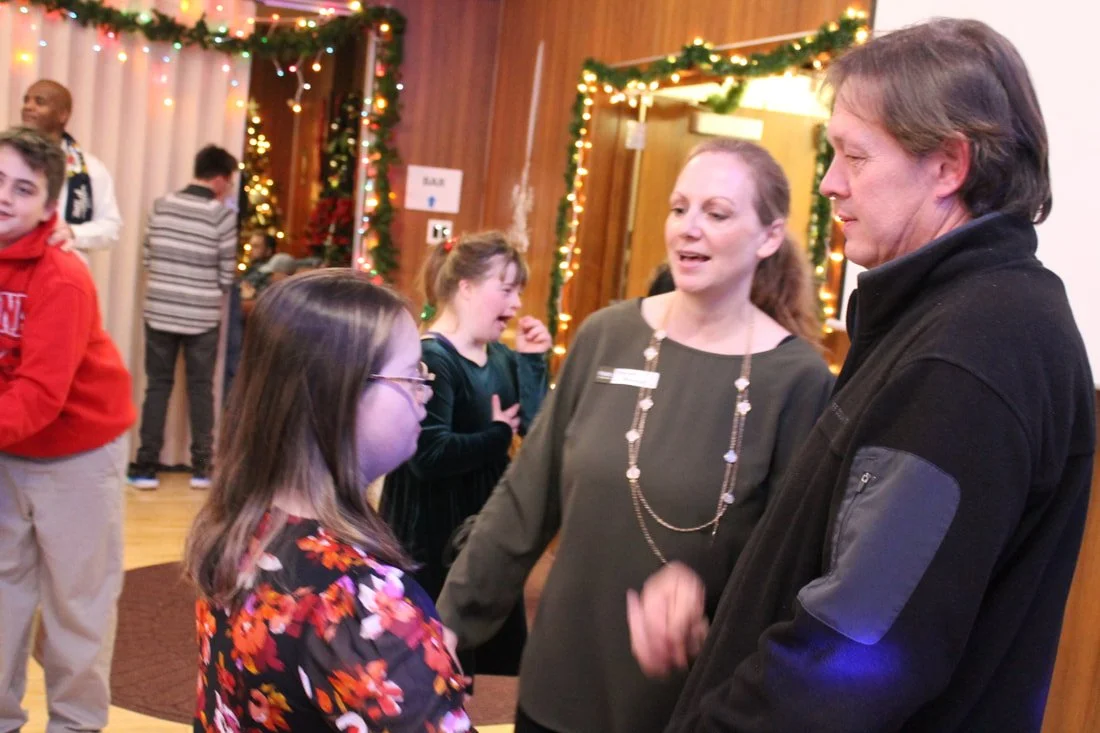A girl with glasses and a floral dress talking to a man and woman at a holiday event, decorated with string lights and greenery.