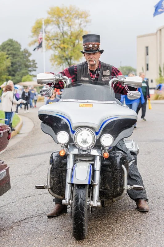 A man wearing a black top hat and a plaid shirt riding a motorcycle at an outdoor event with several people, trees, and flags in the background.