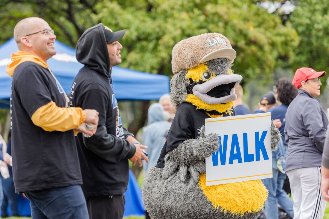 A person in a large bird mascot costume holding a sign that says "WALK" during an outdoor event with people around.