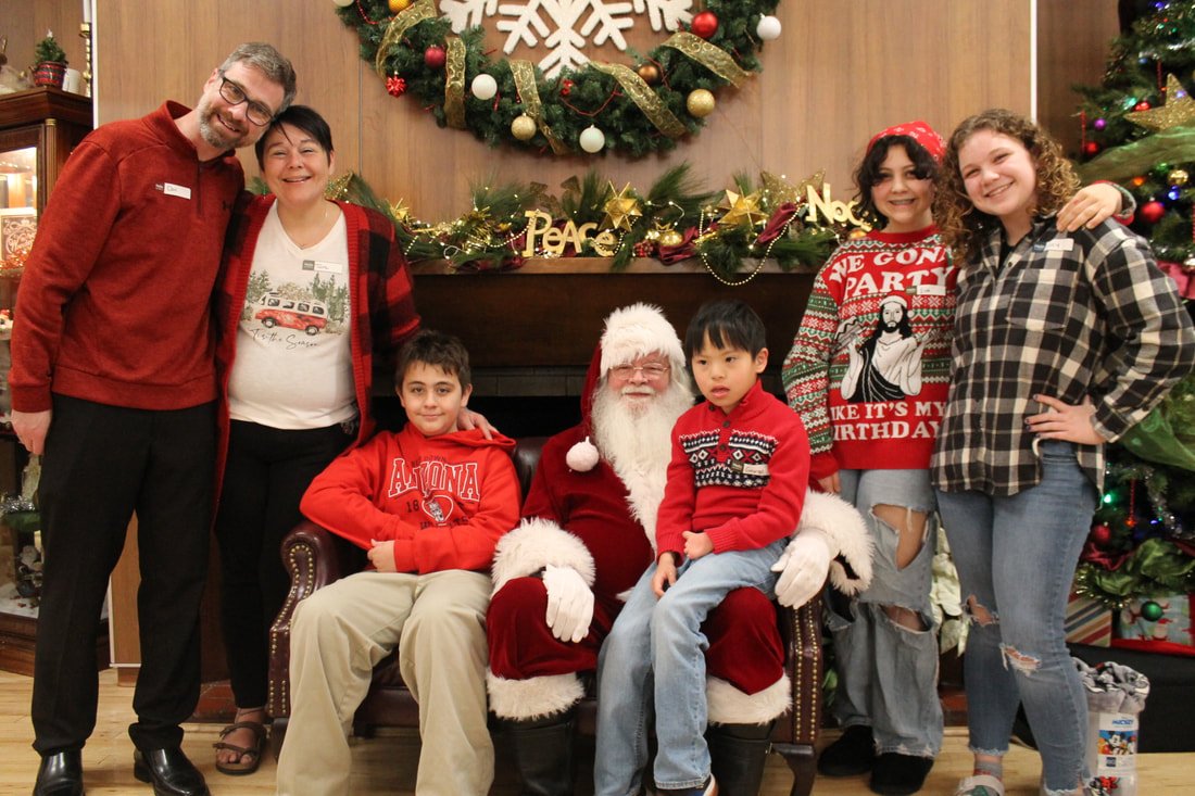 A group of six people, including two children, two women, two men, and Santa Claus, are gathered in front of a Christmas wreath and decorated Christmas tree. The children are sitting on Santa's lap, and everyone is smiling, enjoying a holiday celebra