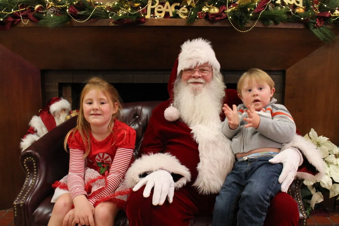 A girl and a boy sitting on Santa Claus's lap during Christmas, with festive decorations and poinsettias in the background.