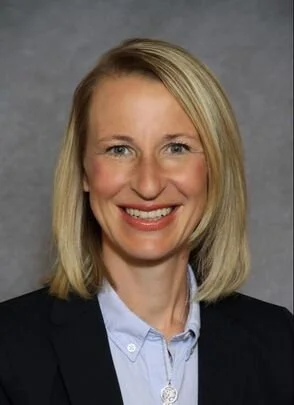 A professional woman with blonde hair and a navy blazer smiling at the camera against a gray background.
