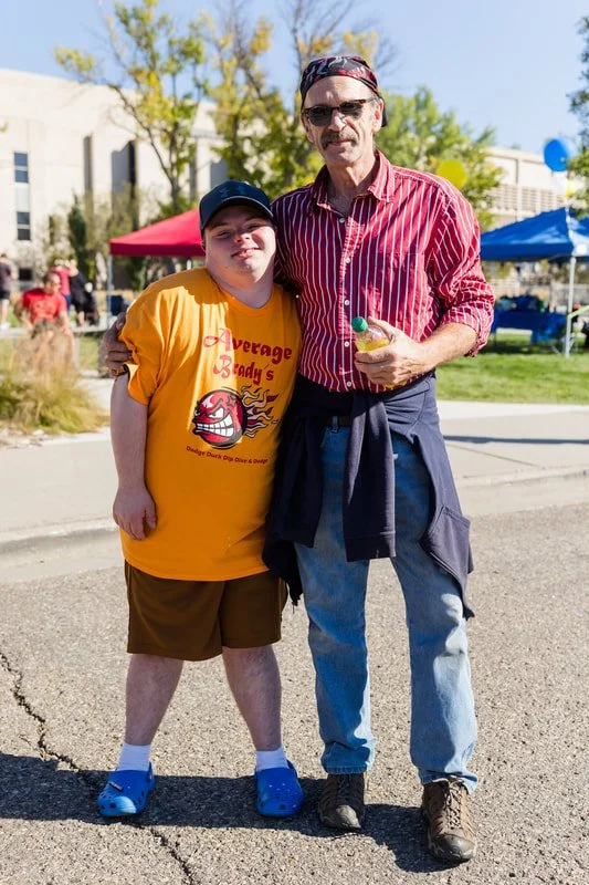 A young boy and an older man smile and pose together at an outdoor event on a sunny day. The boy wears a yellow t-shirt, brown shorts, blue Crocs, and a black cap. The man wears a red striped shirt, light blue jeans, boots, sunglasses, and a headband