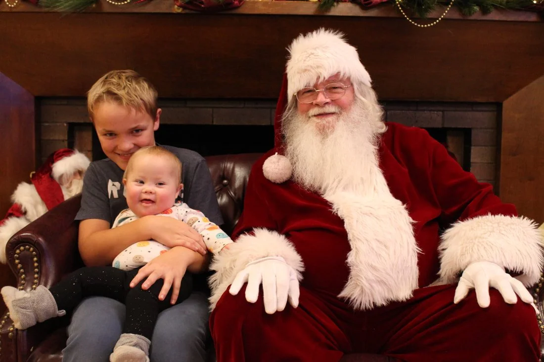 A boy and a baby girl sitting on Santa Claus's lap during Christmas. Santa is smiling, wearing a red suit with white fur trim and glasses. The boy is smiling, and the baby girl is looking happy.