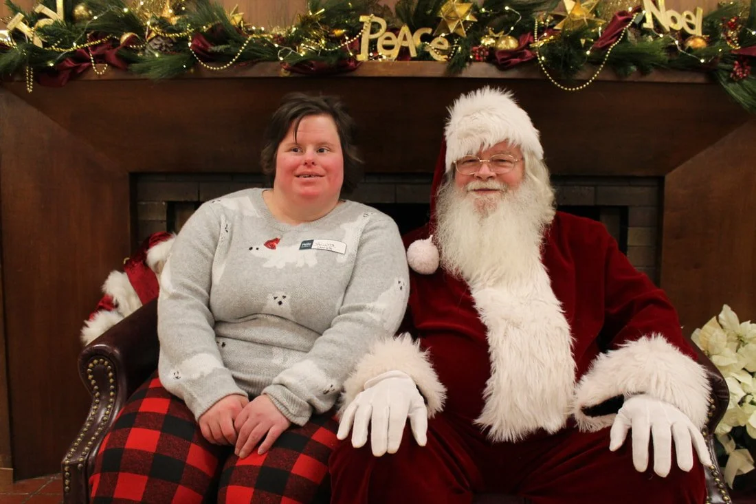 A person with a disabilities sitting next to someone dressed as Santa Claus in front of a decorated fireplace with Christmas garland and ornaments.