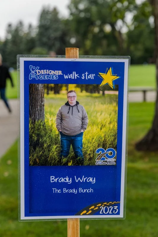 Sign featuring a photo of Brady Wray, a young person in a gray hoodie and glasses, standing outdoors in a grassy field with trees in the background. The sign is blue and includes the text "Designer Genes walk star," the logo for 20 years of Extra Mil