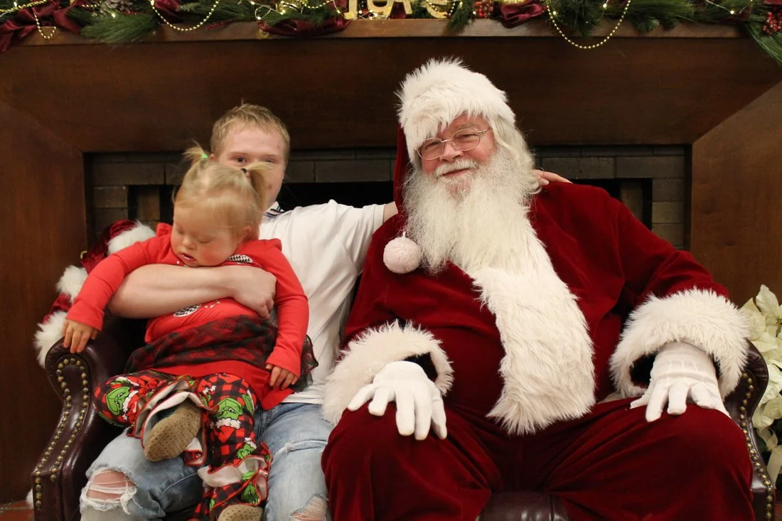 A young man, a little girl, and Santa Claus sitting on a sofa in front of a fireplace decorated for Christmas.