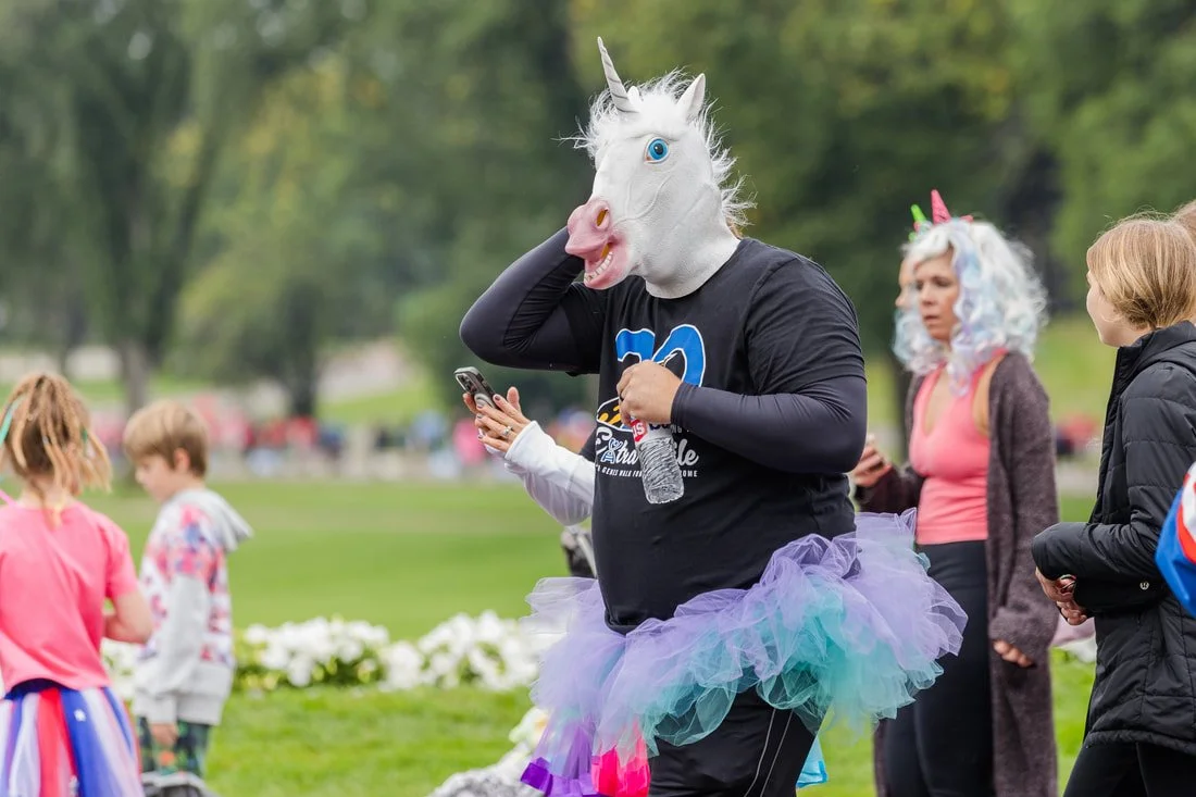 Person wearing a unicorn mask and rainbow tutu at a park during daytime. Several children and adults in costume are nearby.
