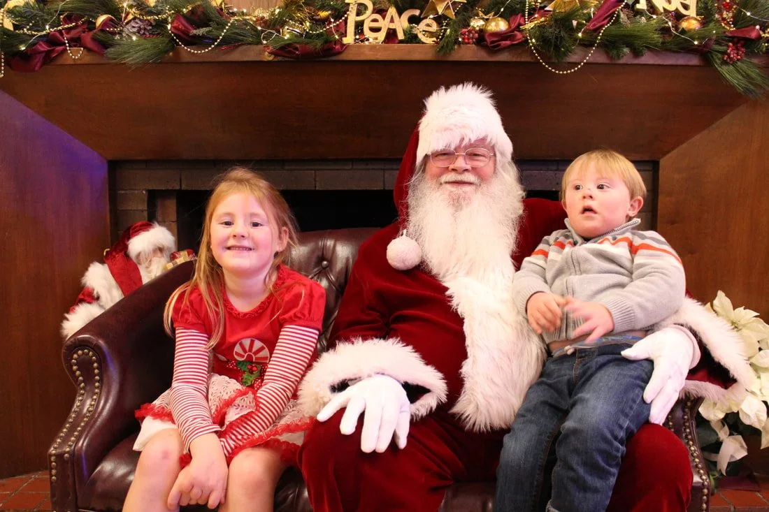 A young girl and a young boy sitting on Santa Claus's lap, with Christmas decorations including garlands, ornaments, and a sign that says 'Peace' in the background.