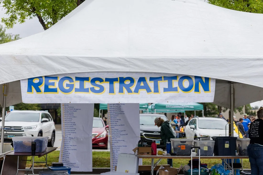 Outdoor registration tent with a large blue and yellow sign reading 'REGISTRATION', with people and cars in the background.