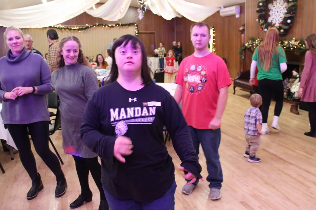 Group of people at a Christmas party in a decorated hall, with Christmas wreaths and lights, some sitting at tables, some standing, and children playing in the background.