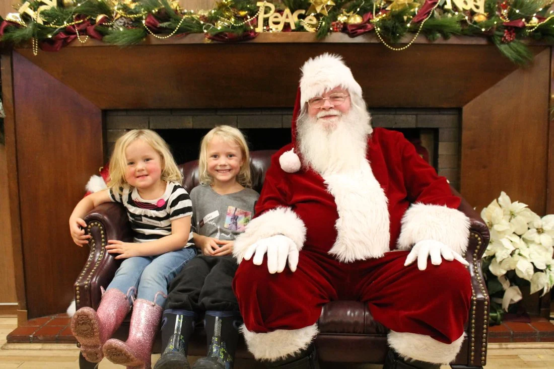 Two young girls sitting beside a man dressed as Santa Claus on a leather sofa in front of a decorated fireplace with Christmas garland and poinsettias.