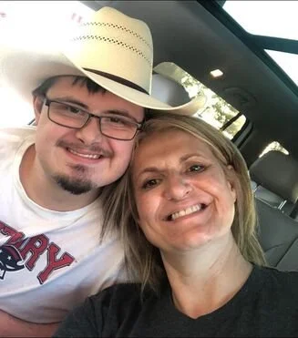 A smiling man and woman taking a selfie inside a car. The man is wearing glasses and a cowboy hat, and the woman has blonde hair.