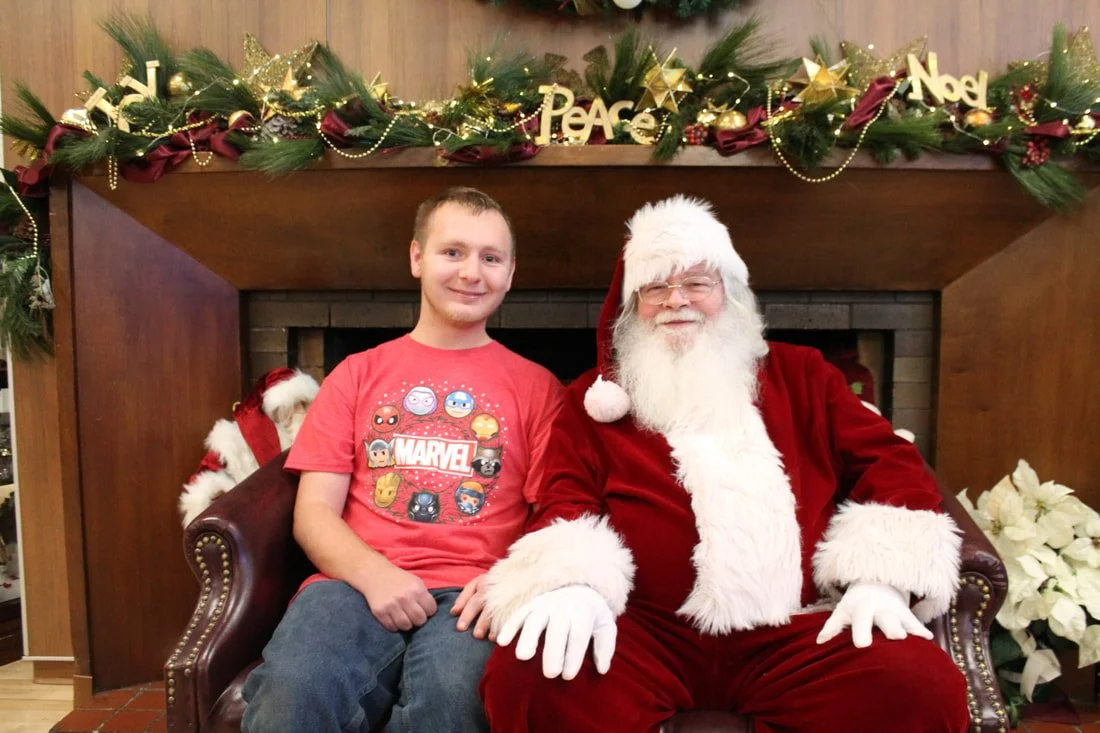 A young man sitting next to Santa Claus on a holiday-themed sofa. The background features Christmas decorations, including garland, ornaments, and the words "Peace" and "Noel."