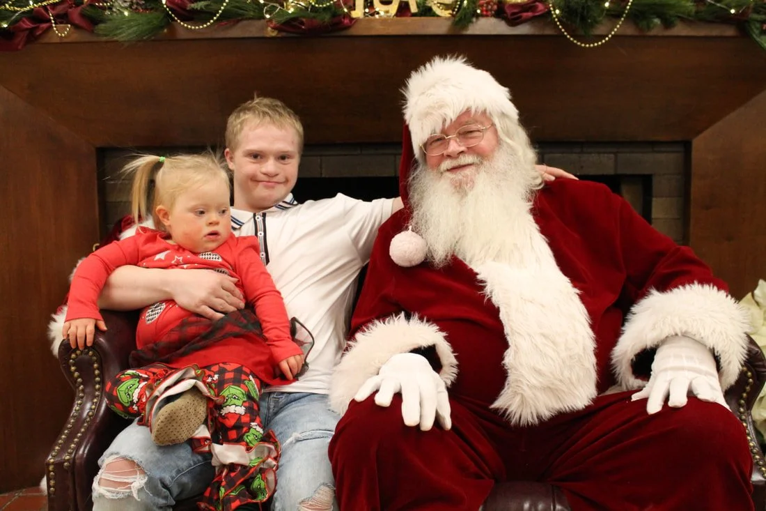 Three people sitting with Santa Claus for a holiday photo. The man dressed as Santa has a white beard, glasses, and a red suit with white fur trim. The boy has blond hair, a white shirt, and ripped jeans. The girl, sitting on the boy's lap, is wearin