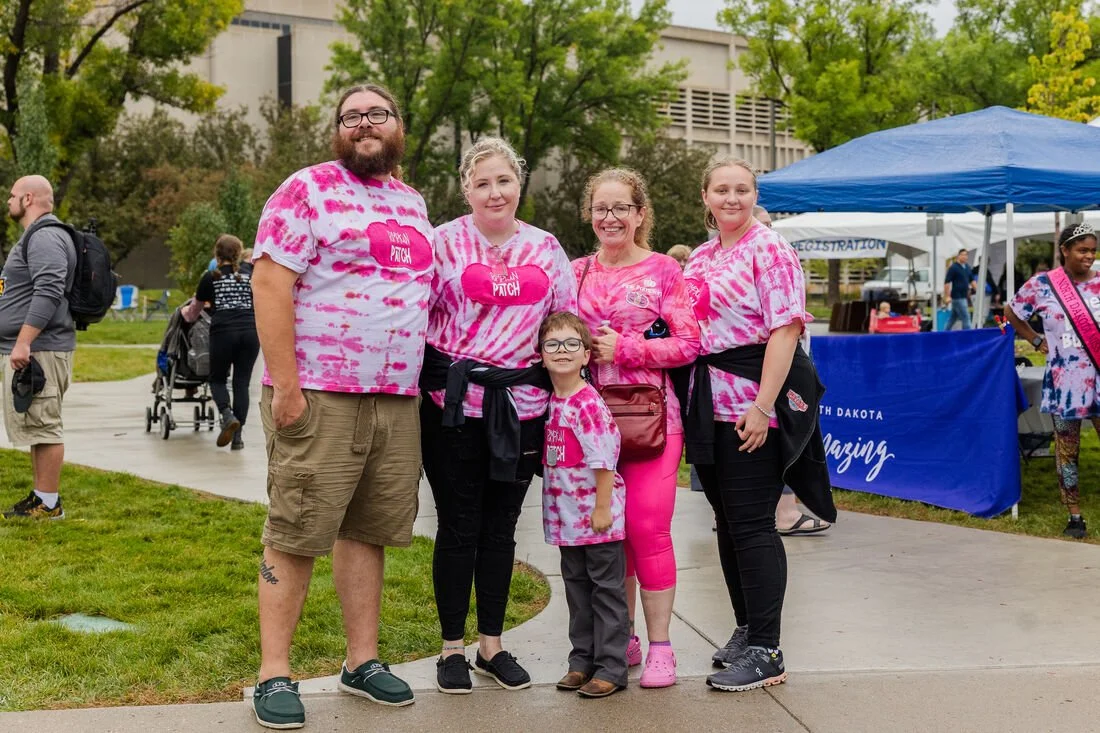 Group of five people, four women and one girl, wearing matching pink tie-dye shirts at an outdoor event during daytime. They are standing on a sidewalk with green trees and event tents in the background.