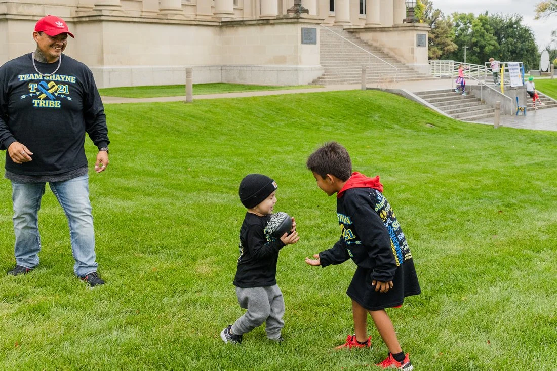 Two children, one younger and one older, are playing with a football on a grassy area in front of a large building. An adult man is nearby, smiling and watching them. The children are wearing dark clothing with hoodies, and the younger child is also 