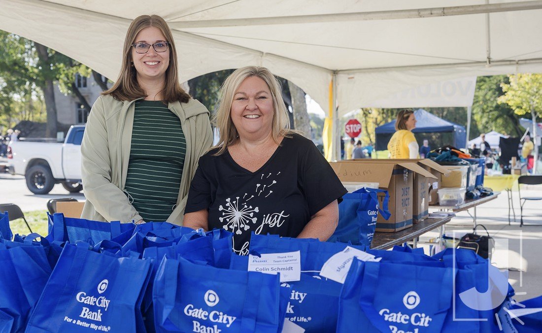 Two women smiling at a booth with blue bags under a white tent at an outdoor event.