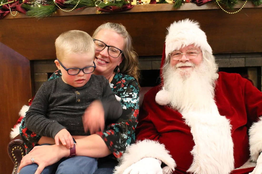 A woman holding a young boy sitting next to a man dressed as Santa Claus, all smiling, at a Christmas event.