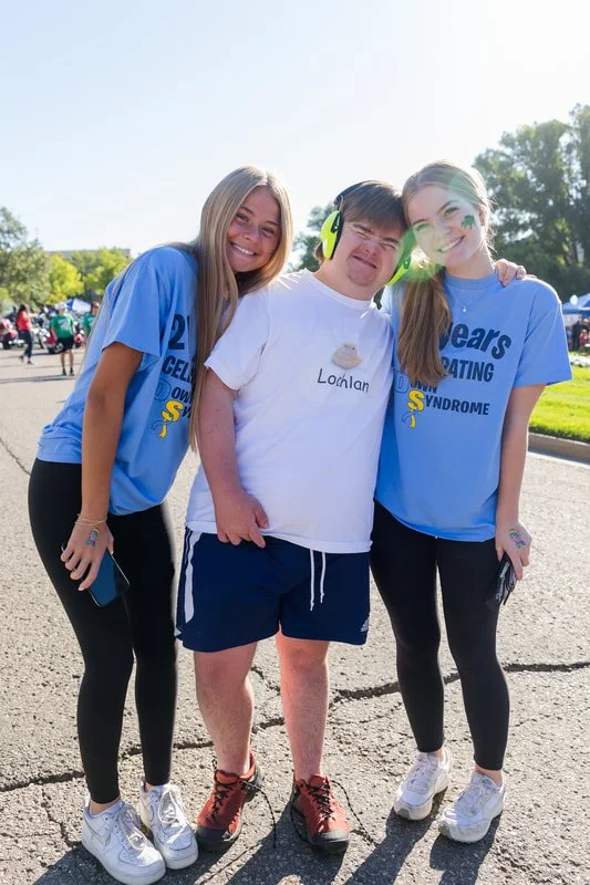 Three people smiling and hugging at an outdoor event, two young women in blue T-shirts and a young man in a white T-shirt with hearing protection and a hat, on a sunny day.