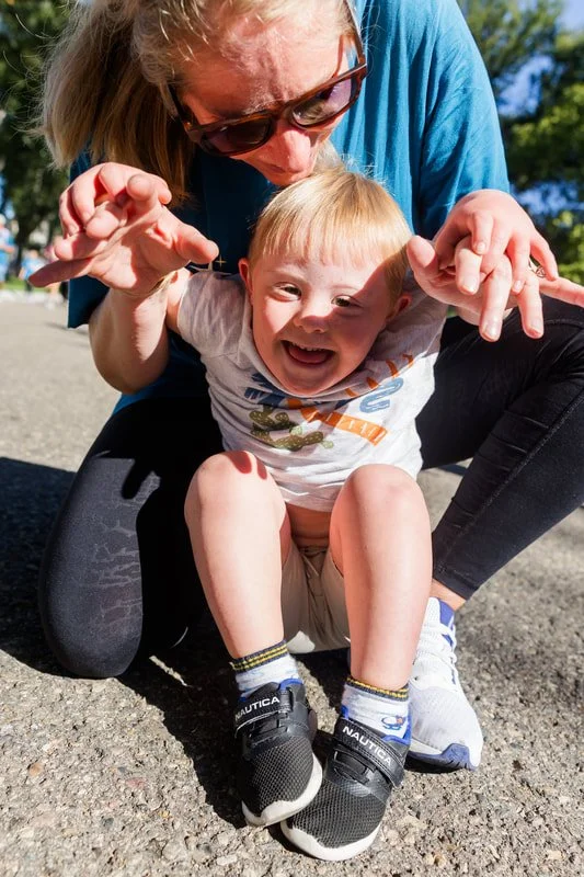 A woman playfully holds a young child's arms, and they are both smiling and laughing outdoors on a sunny day.