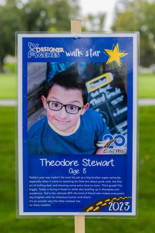 Signboard honoring Theodore Stewart, 8 years old, with a portrait of a young boy with glasses and short dark hair. The sign features a blue background with a yellow star, a logo for Designer Genes Walk Star, and a 20th anniversary emblem for Extra Mi