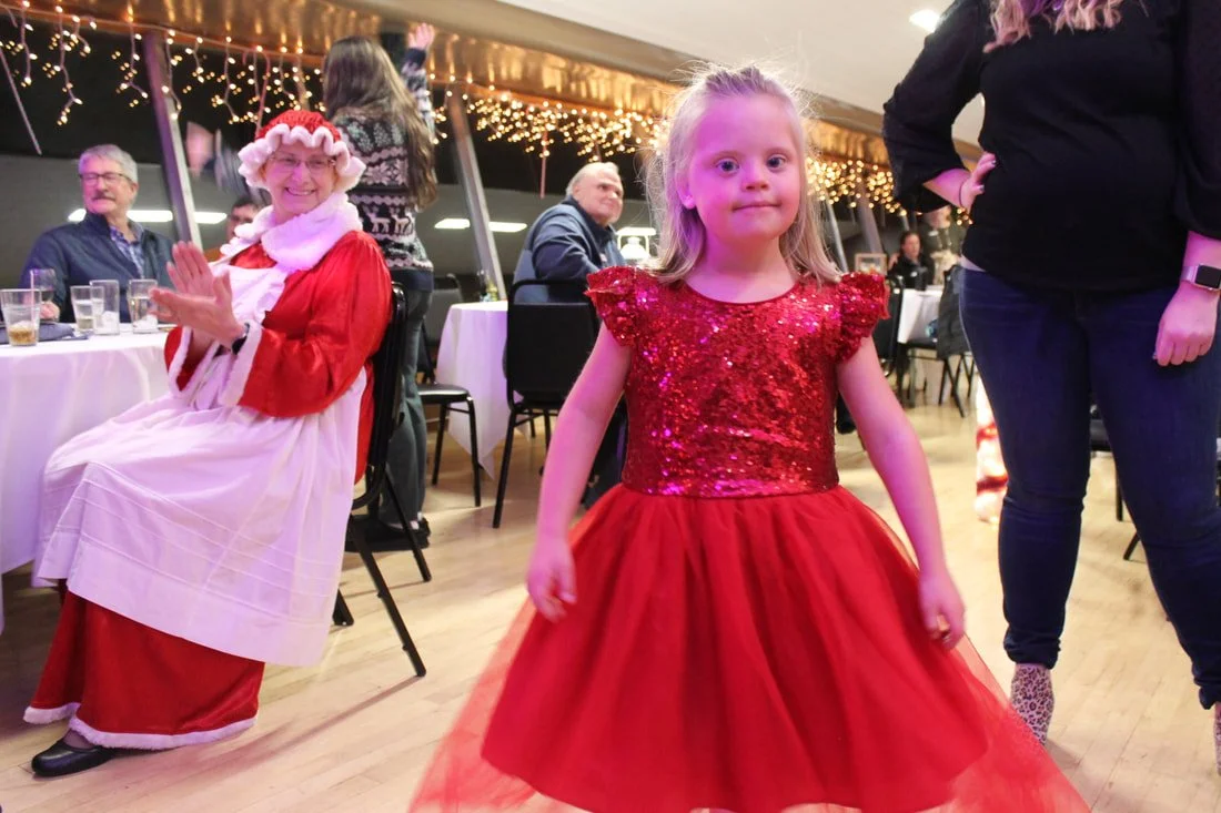 A young girl in a red, sparkly dress standing in a decorated party room with Christmas lights, surrounded by seated adults and standing people, including a woman dressed as Santa Claus.
