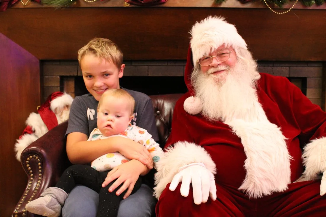 A smiling boy sitting on a brown leather chair holding a baby, next to Santa Claus who is dressed in a red suit with white fur trim, wearing glasses, and smiling. There is a Christmas stocking hanging on the fireplace behind them.