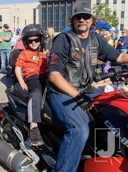 A man and a young girl sitting on a motorcycle at a public event, with people gathered in the background. The man is wearing sunglasses, a cap, a leather vest, and jeans, while the girl is wearing a helmet, sunglasses, and a red shirt.