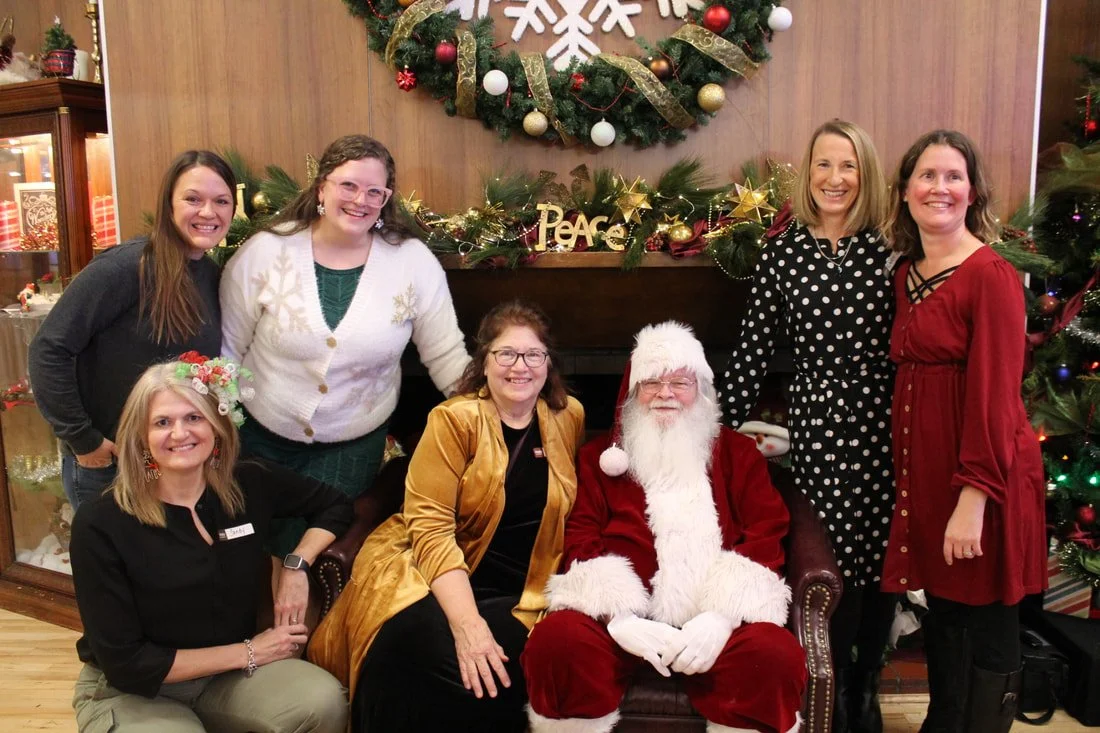 Group of eight women and Santa Claus sitting and standing in front of a Christmas decorated fireplace with a large Christmas wreath and presents.