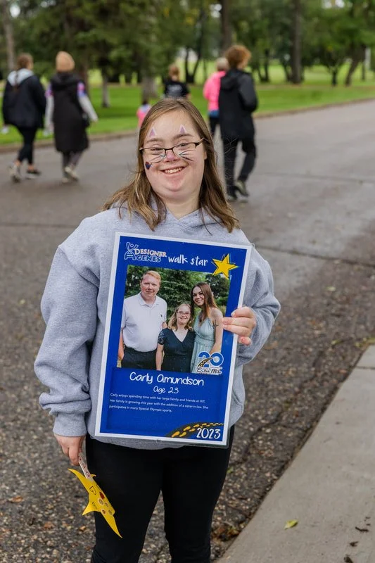 Young girl at a park holding a certificate celebrating her participation in the Special Olympics, surrounded by trees and other people in the background.
