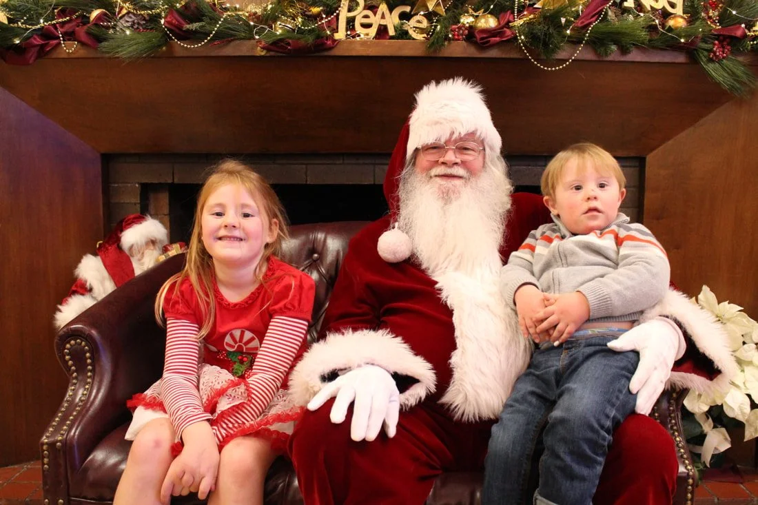 A girl and a boy sitting on Santa Claus's lap inside a decorated fireplace area for Christmas. The girl is smiling, wearing a red dress with Christmas decorations on it and striped sleeves. The boy looks surprised, wearing a gray hoodie with orange a