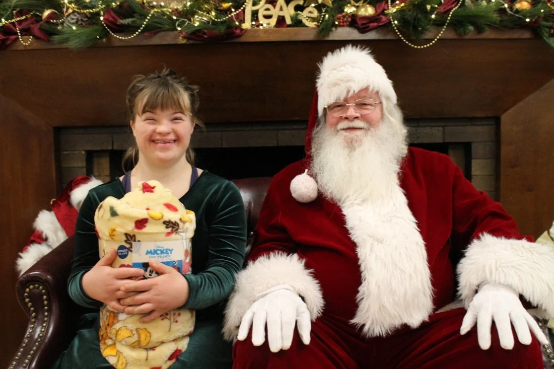 A young girl happily sits next to Santa Claus, holding a colorful plush blanket, in a festive holiday setting with Christmas decorations and greenery in the background.