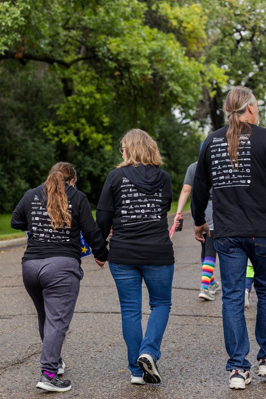 Three women walking hand in hand outdoors during daytime, wearing matching black hoodies with logos and text on the back, on a paved path surrounded by green trees.