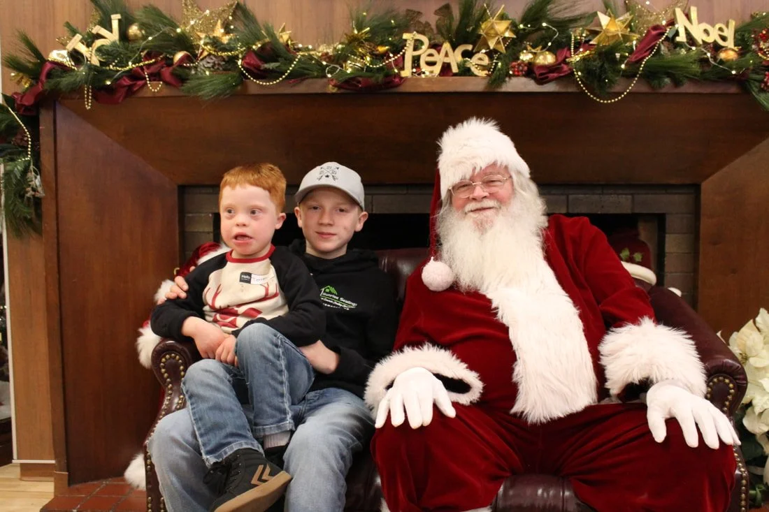 Two children sitting on Santa Claus's lap in front of a decorated fireplace with Christmas ornaments.