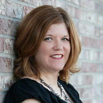 Portrait of a woman with shoulder-length light brown hair, smiling slightly, standing against a brick wall.