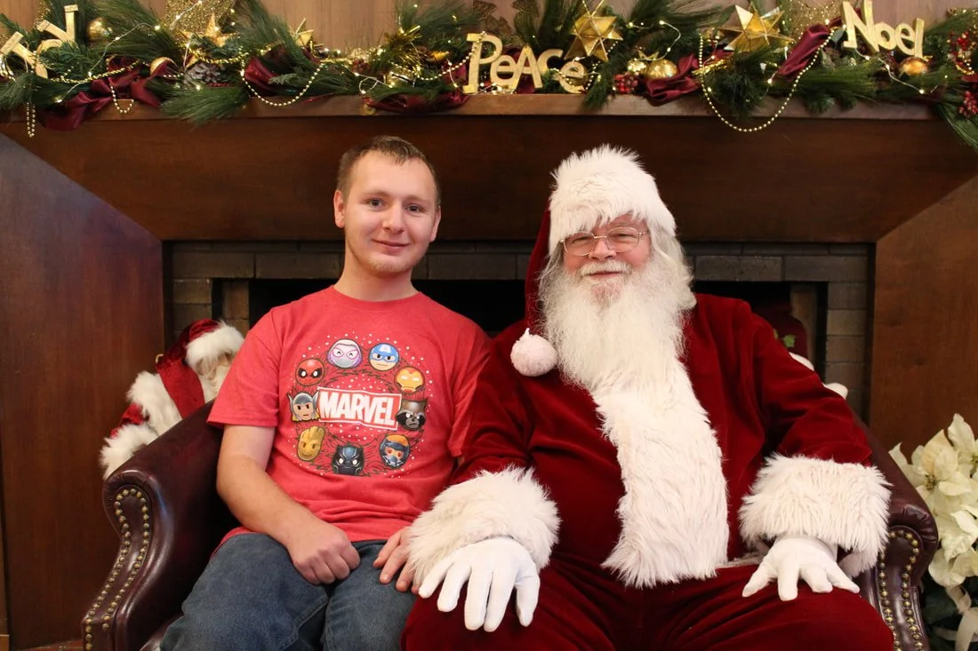 A young man sitting next to Santa Claus, both smiling, in front of a decorated fireplace with Christmas ornaments and greenery.