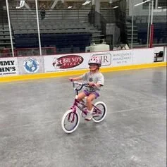 A young girl riding a pink and white bicycle on an ice rink, wearing a helmet and casual clothes.