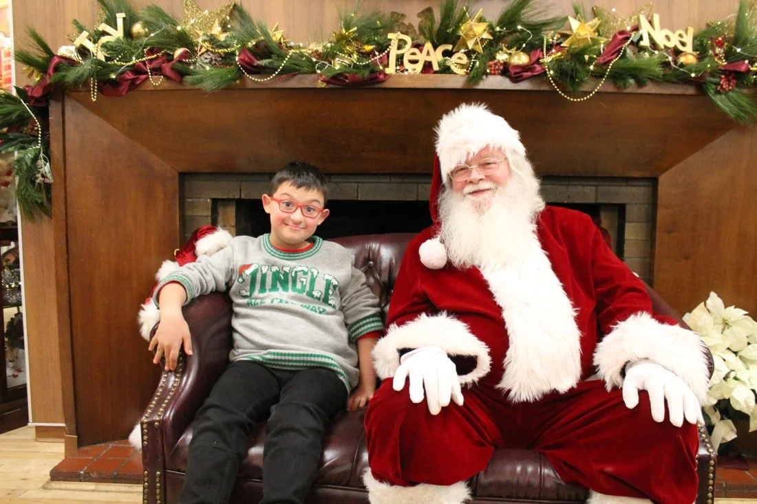 A young boy sitting next to Santa Claus on a couch, smiling. The setting is decorated with Christmas garland, ornaments, and the words 'Peace' and 'Noel' in gold above the fireplace.