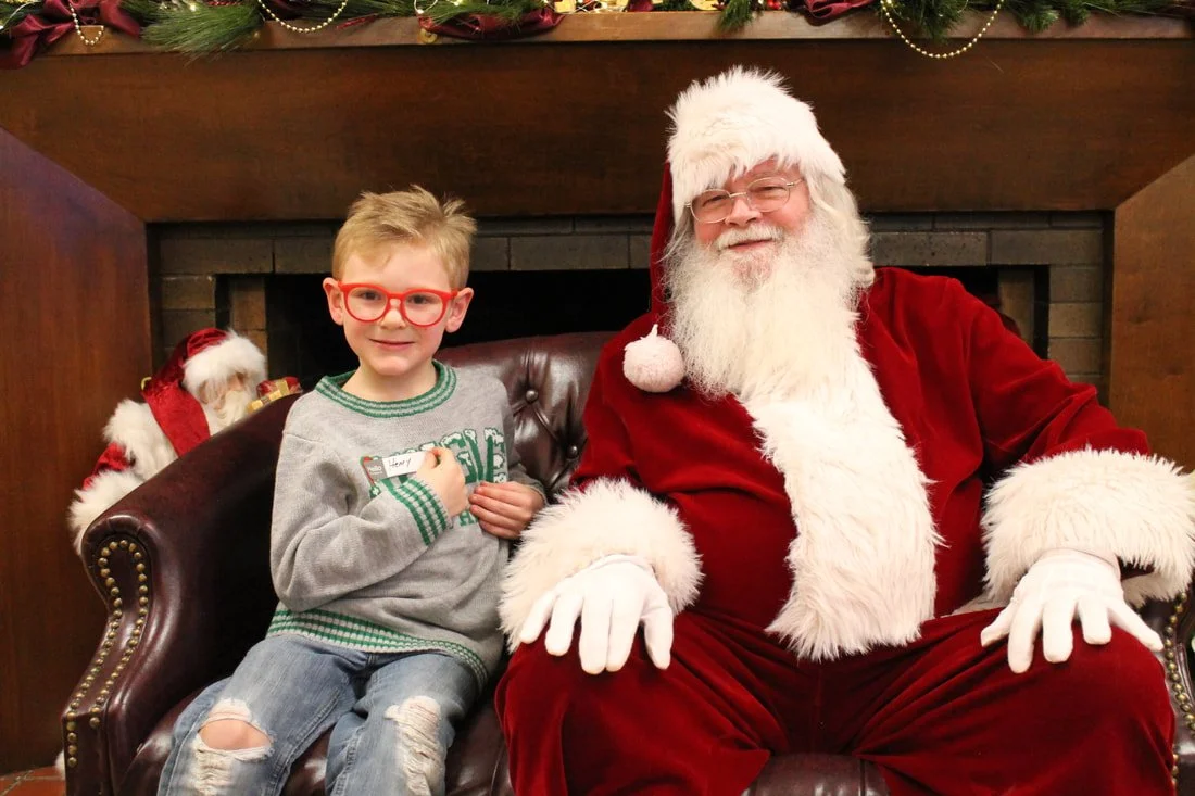 A young boy with glasses sitting next to Santa Claus in a festive setting, both smiling.