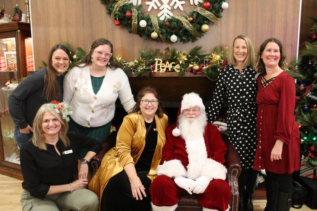 Group of six women and a man dressed as Santa Claus gathered in front of a decorated Christmas fireplace, smiling. The background features a holiday wreath, Christmas ornaments, and decorations.