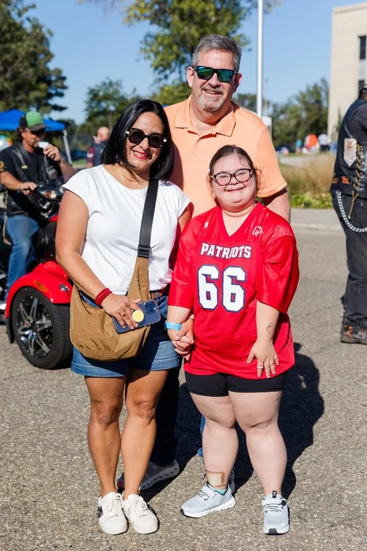 Three people standing outdoors on a sunny day, posing for a photo, with a motorcycle and other people in the background. The woman on the left has shoulder-length black hair, sunglasses, a white t-shirt, denim shorts, and white sneakers. The man in t